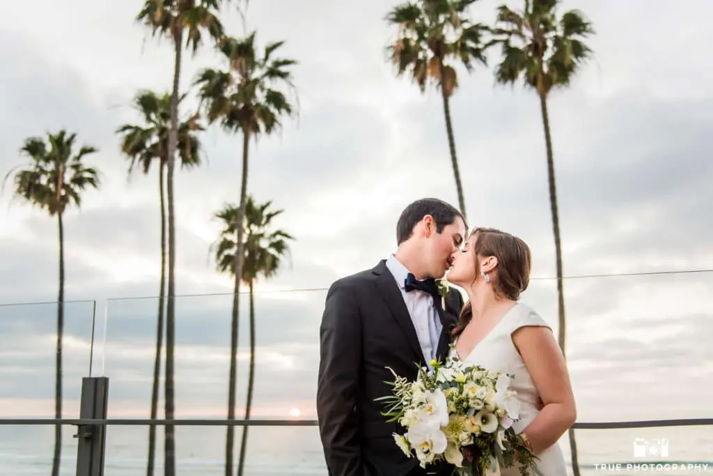 The newlyweds enjoy a private kiss on the balcony, palm trees behind them swaying in the ocean breeze.
