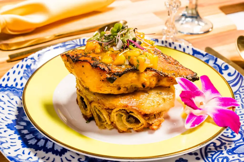 Grilled fish fillet topped with tropical salsa, served on a layer of rolled delicacies, presented on a decorative blue-patterned plate
