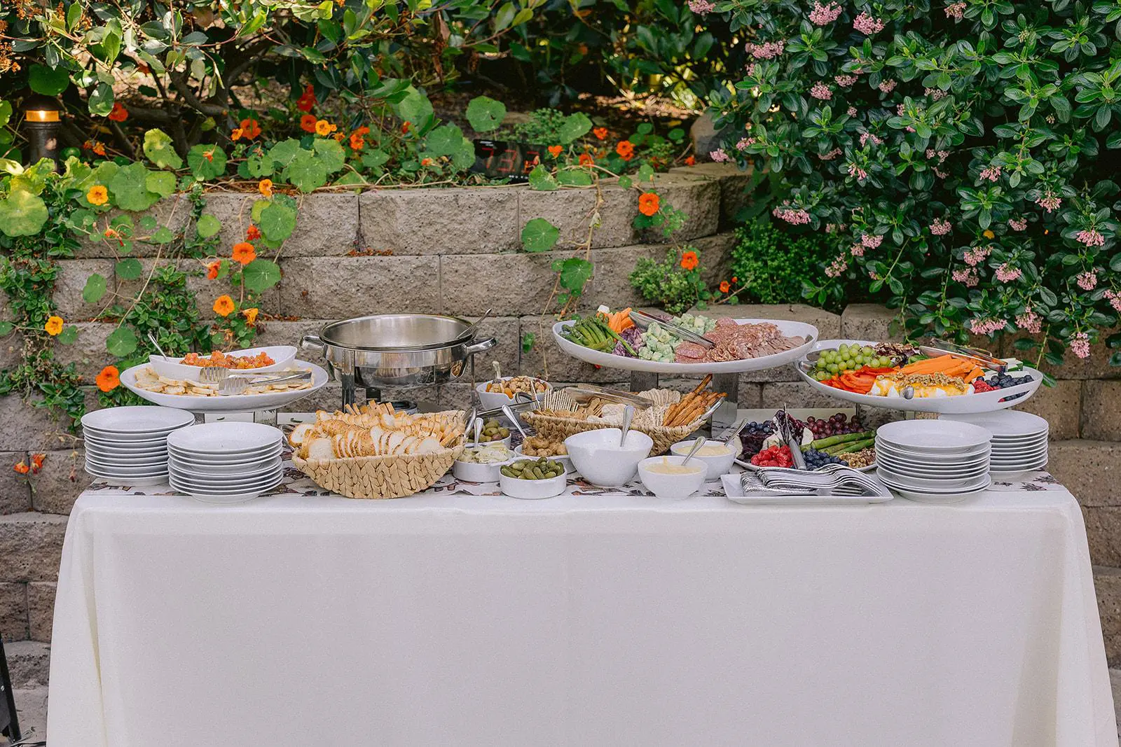 Buffet spread featuring fresh vegetables, fruits, crackers, and dips arranged neatly on a long table covered with white cloth outdoors