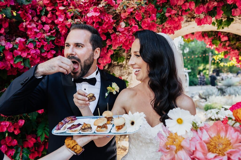 Bride and groom enjoying hors d'oeuvres outdoors, surrounded by vibrant red flowers, with the groom tasting a treat while the bride smiles joyfully, both adorned with floral details