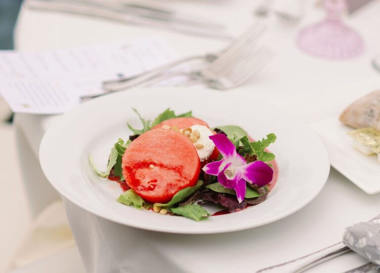 Plated dish from San Diego wedding catering: Fresh salad with a vibrant red slice of tomato, accented by a purple orchid, on a white table setting