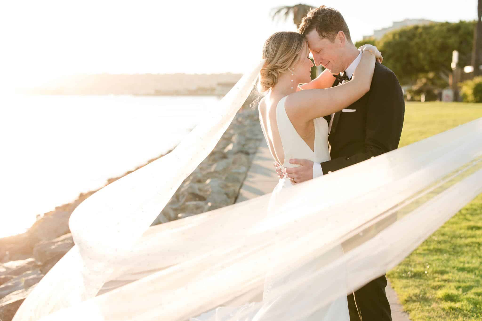 Bride and groom sharing a romantic embrace with flowing veil in the sunset by the seaside