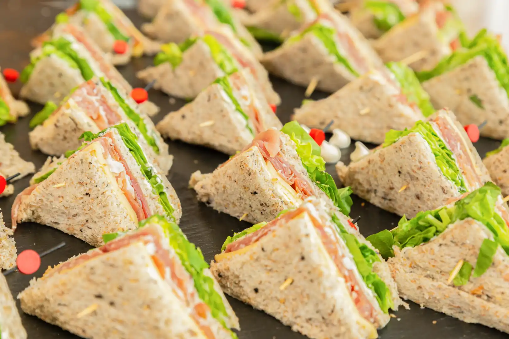 Close-up of an array of triangular whole wheat sandwiches arranged on a dark platter for catering, filled with lettuce, cheese, and deli meat, held together with toothpicks and red decorative picks.