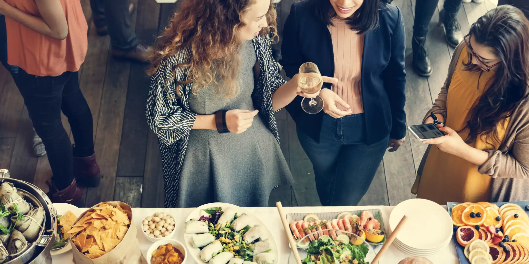 High-angle shot of three women standing around a catered buffet table with shrimp, chips, salad, and fresh fruit; one holds a wine glass while chatting and another checks her smartphone during a graduation party.