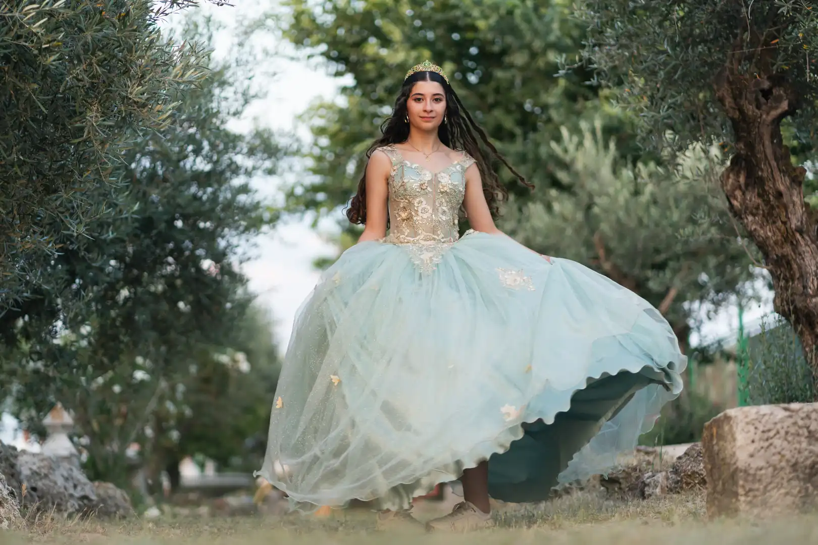 Elegant young woman posing in a tiara and elaborate light green quinceañera gown for an outdoor portrait session.